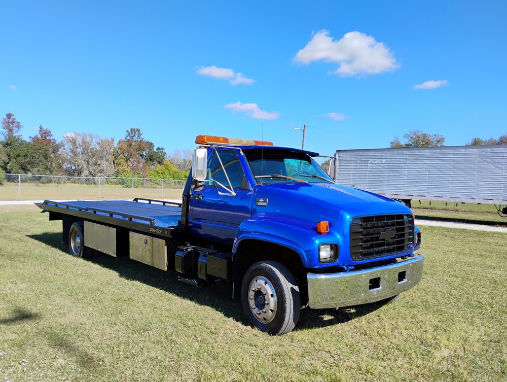 Blue flatbed tow truck parked on grass with trees and buildings in background