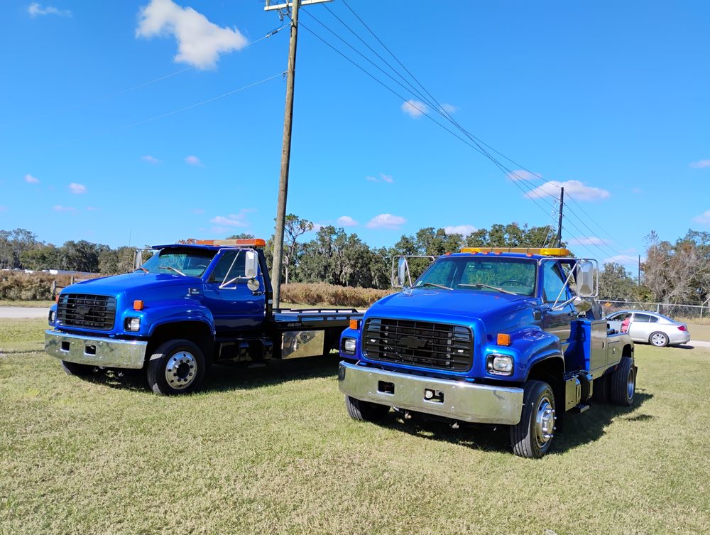 Two blue and tan vintage tow trucks parked on grass with utility poles and trees in background