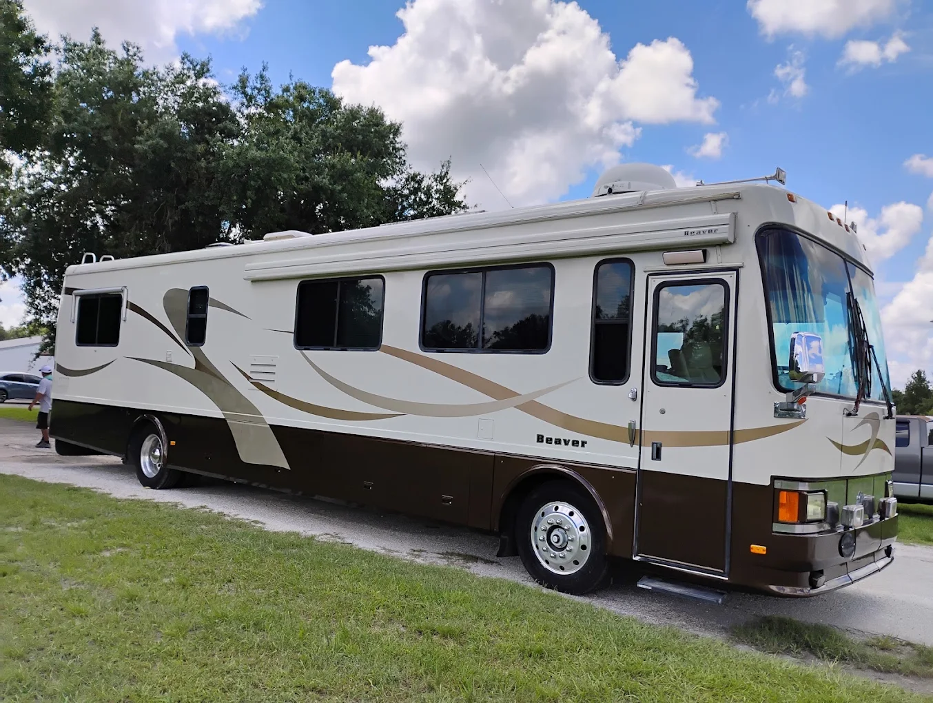 White and tan Beaver motorhome parked on grass with trees and blue sky in background