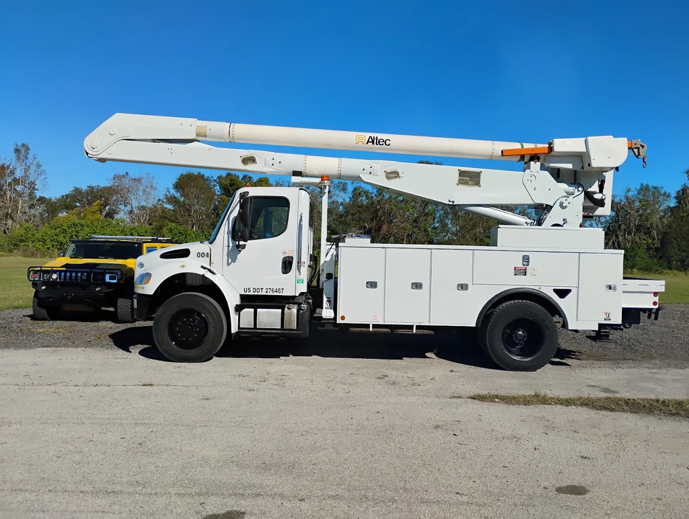 White Altec bucket truck with extended boom parked on gravel lot with trees in background