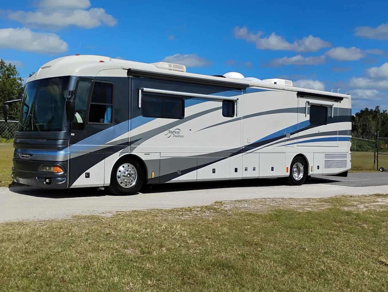 White and blue motorhome parked on grass with clear sky and trees in background
