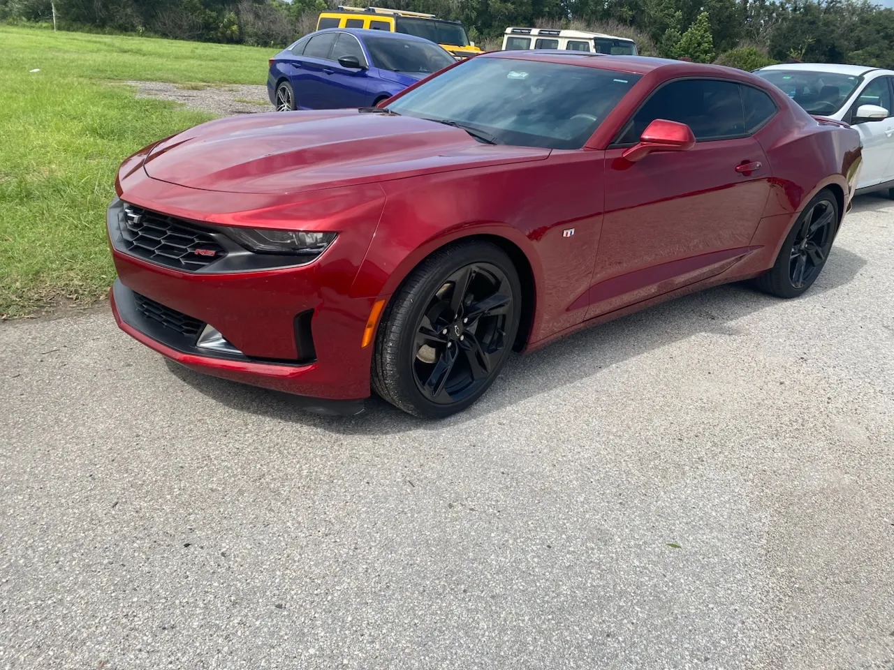 Red Chevrolet Camaro sports car parked on asphalt lot with green grass and vehicles in background