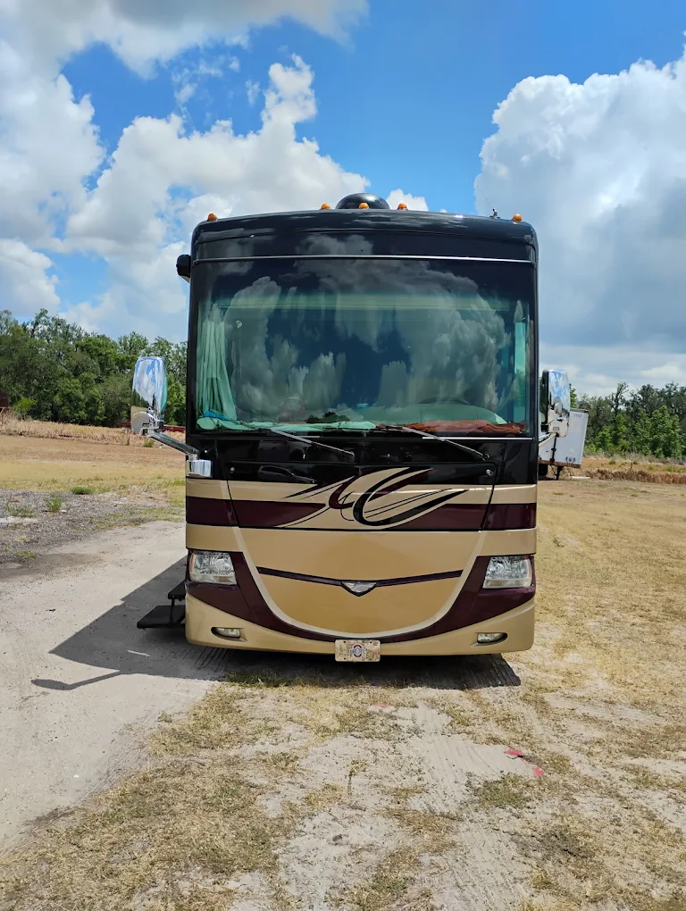 Beige and burgundy motorhome parked on dirt lot under blue sky with white clouds