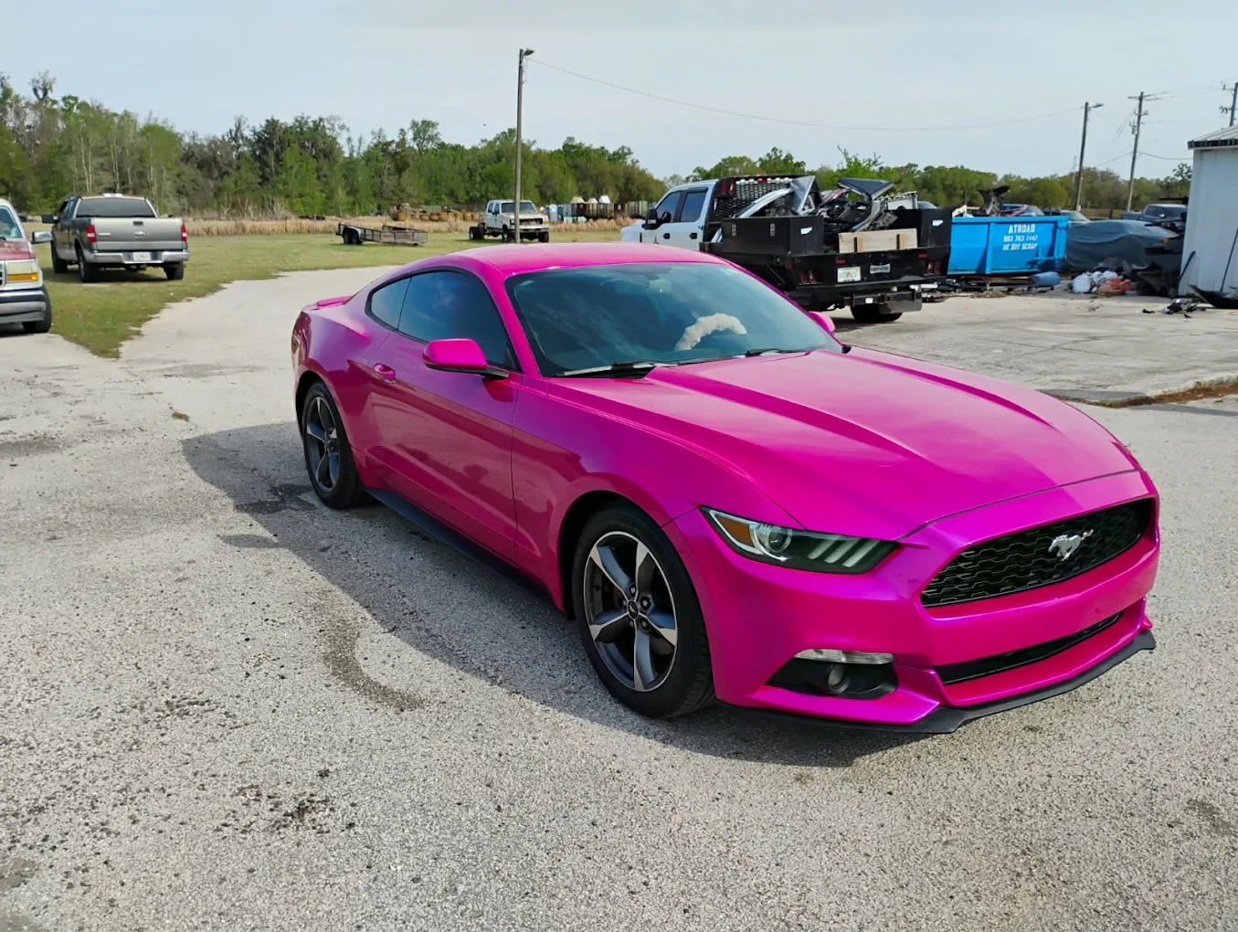 Bright pink Ford Mustang sports car parked in lot with trucks and buildings in background