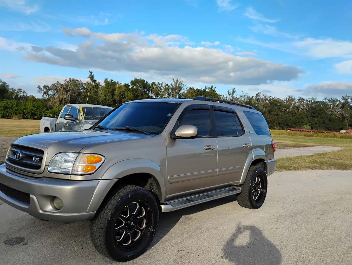 Silver Toyota Sequoia SUV with custom wheels parked on asphalt, white truck visible in background