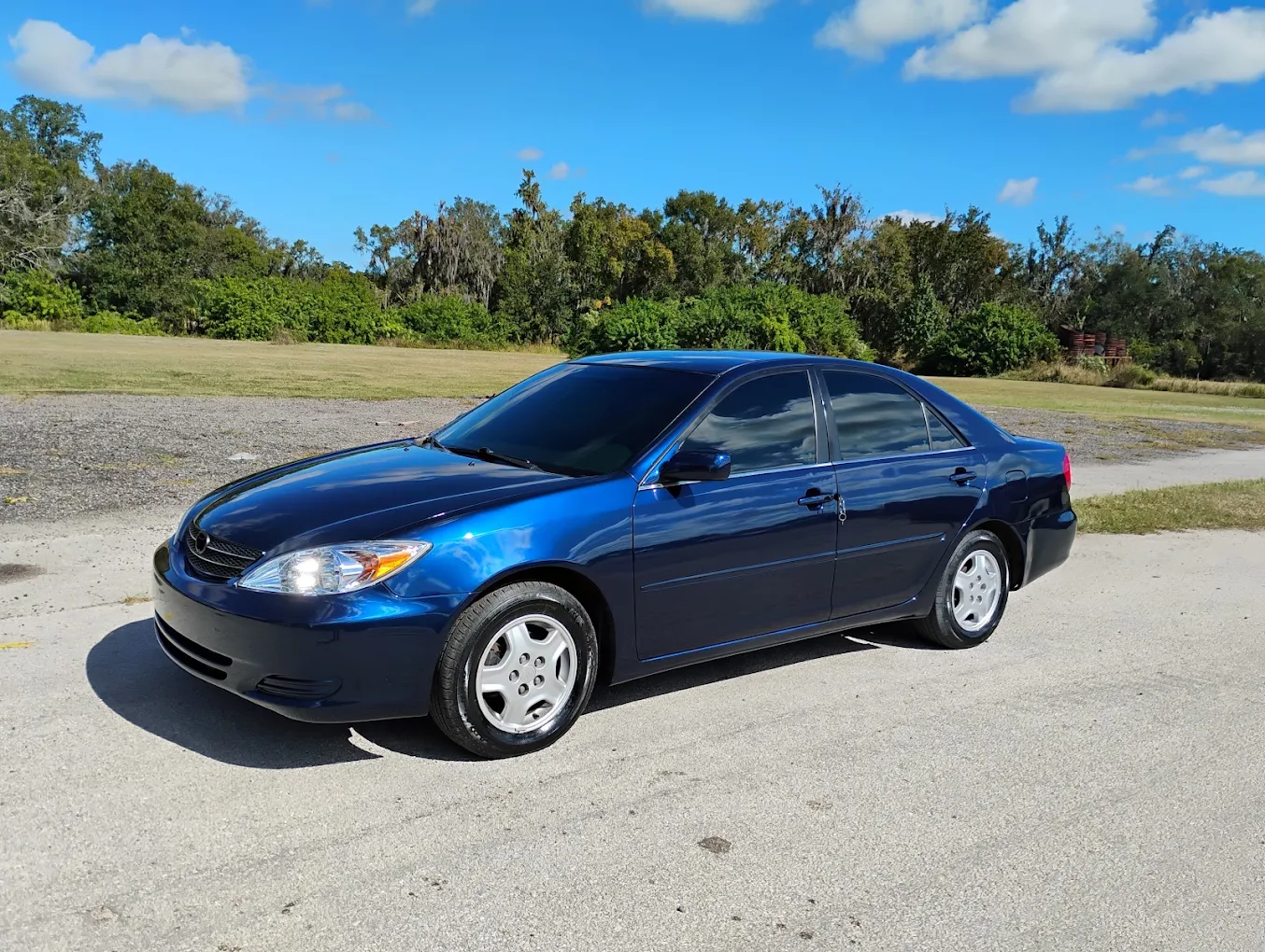 Blue Toyota Camry sedan parked on gravel driveway with trees and clear sky in background